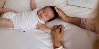 An image of a newborn baby holding the hand of a female support person. She is gently patting the newborn on the head while he has his eyes closed wearing a singlet and a nappy.