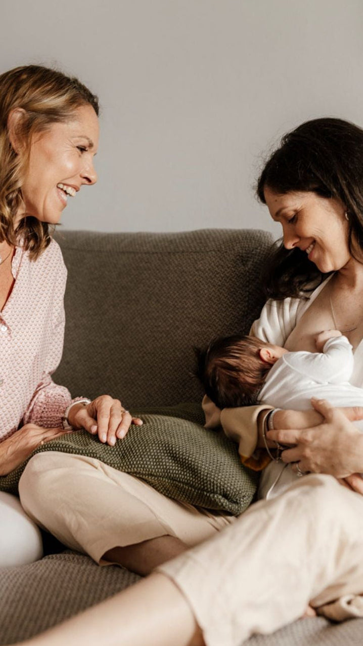 A woman in a pink patterned top and beige pants sits on a gray couch beside another woman holding a baby wrapped in white. The room has a soft neutral color palette with a gray wall and textured sofa.